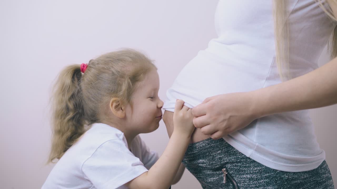 귀여운 작은 소녀가 임신한 어머니의 배를 밝은 방에서 키스합니다.