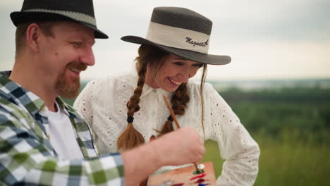 an artist, holding a paint palette, working on a large canvas in an outdoor setting. beside him, a woman in a white dress and hat stands, smiling joyfully as she observes his work