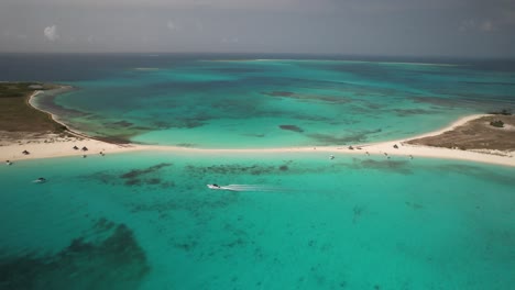 Ein-Boot,-Das-Durch-Türkisfarbenes-Wasser-In-Der-Nähe-Eines-Sandstrandstreifens-Bei-Los-Roques-Navigiert,-Luftaufnahme