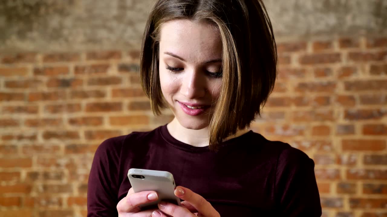 Young cute girl is reading message on her smartphone, she is surprised, communication concept, brick background
