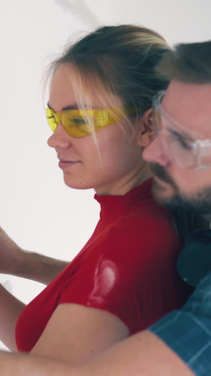 CU, Dolly cam: young woman in goggles with boyfriend checks level of white wall in empty light renewing room closeup