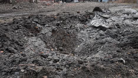 tilt-down view of a deep missile crater is seen in the foreground of a muddy street in a ukrainian village. in the background, the destroyed roofs of houses show the impact of the blast