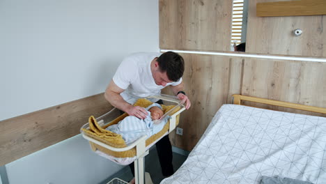 caucasian father stands near the crib with his newborn child. daddy kisses his sleeping baby. parent and infant in after birth hospital ward.