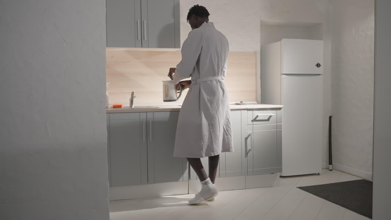 Man in white bathrobe standing in modern kitchen, preparing breakfast or looking at counter. Bright lighting illuminates kitchen space with minimalist design. Clean, tidy kitchen environment