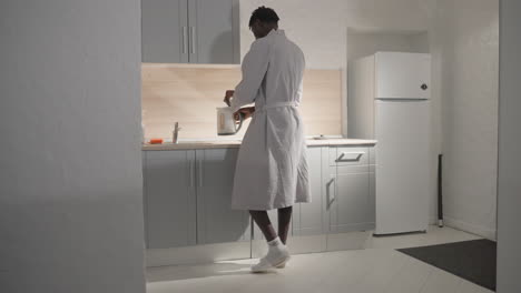man in white bathrobe standing in modern kitchen, preparing breakfast or looking at counter. bright lighting illuminates kitchen space with minimalist design. clean, tidy kitchen environment