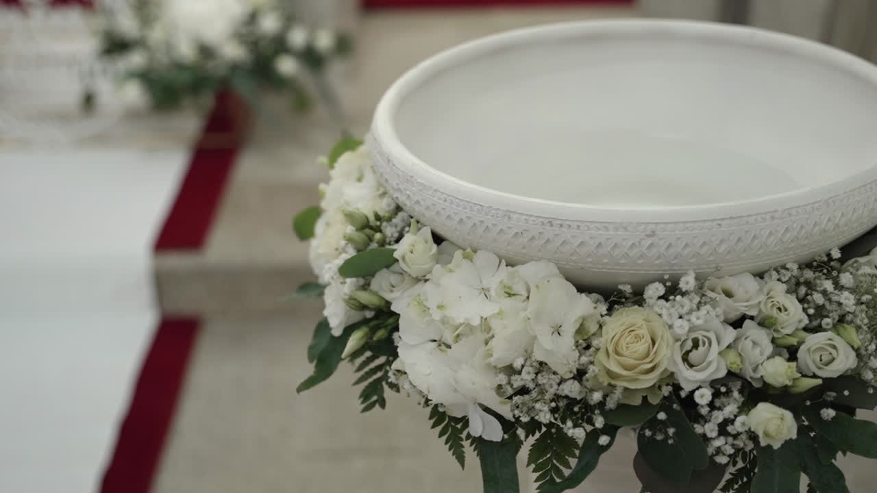 Ornate baptismal font surrounded by delicate white roses, hydrangeas, and lush greenery