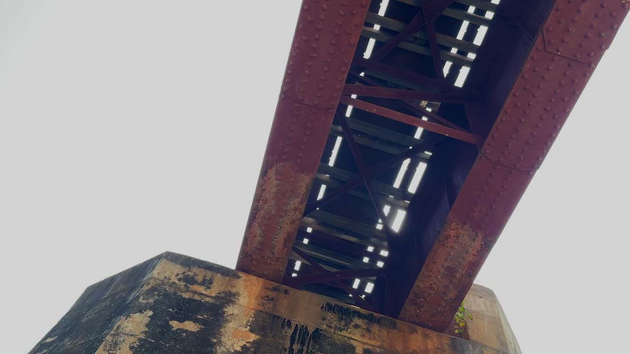 Panorering under en rusten jernbanebro, der afslører forvitrede bjælker og støttepiller, mens kameraet bevæger sig langs konstruktionen mod en lys himmel