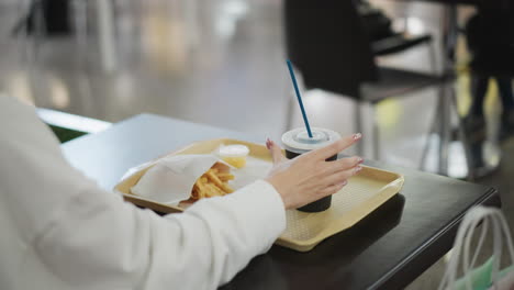 hand inserting straw into coffee cup placed on tray takes it up, with covered fries, set on table in casual dining setting with chairs and soft blurred background