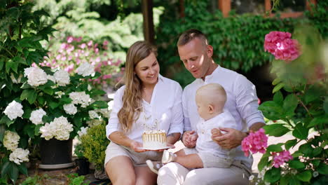 beautiful caucasian couple sit outdoors surrounded by lush greenery and flowers. man holds a cute baby trying to reach a cake in mom's hands.