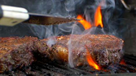 Eine-Fleischzange-Schiebt-Ein-Fast-Gekochtes,-Saftiges-Rib-Eye-Steak-Auf-Einen-Grill-Und-Drückt-Darauf,-Während-Flammen-Emporschießen