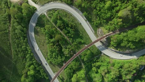 static aerial overhead footage of multiple cars driving on a winding road intersected by an old railway bridge in the middle of a forest