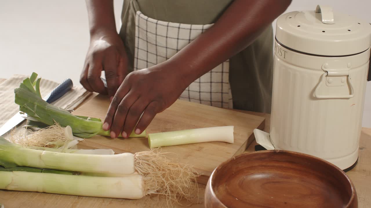Uomo afroamericano che prende un porro, taglia le radici e lo affetta con un coltello sul piano di lavoro della cucina per cucinare.
