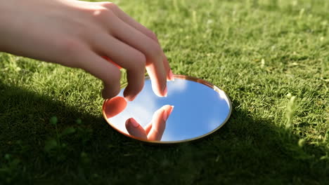 hand reaching for a mirror reflecting the sky on grass