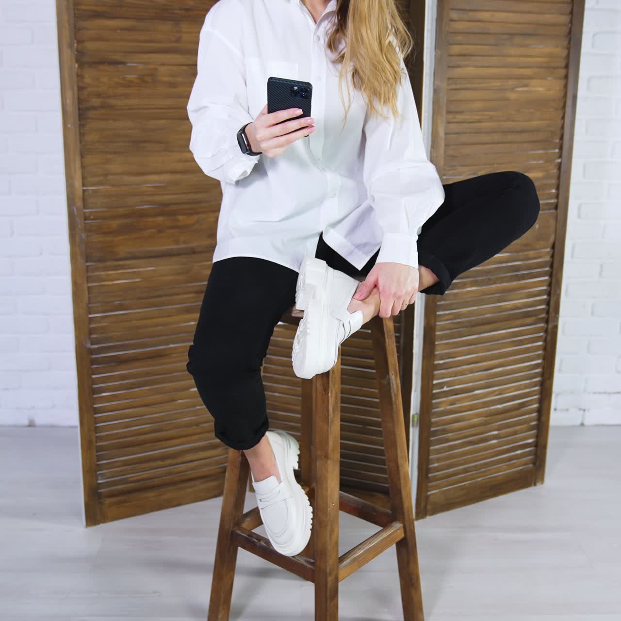 Young long-haired woman sits on wooden stool showing white shoes. Lady in shirt and trousers demonstrates a pair of shoes and uses phone