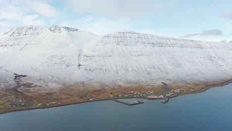volando hacia una pequeña ciudad portuaria en el mar en los fiordos del oeste, islandia con un muelle - magníficos acantilados oceánicos de montaña cubiertos de nieve blanca, antena de drones sobre aguas árticas