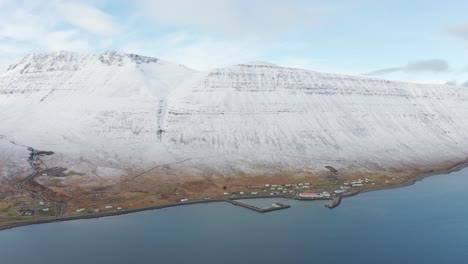 flying towards small harbor town at sea in westfjords, iceland with boat dock - magnificent white snow covered mountain ocean cliffs, drone aerial over arctic waters