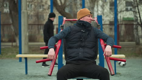 a coach seated on workout equipment outdoors, resting his hands on red rods, looks tired as he looks to the left, in the background, a boy dressed in black plays football
