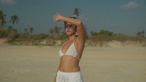 A-young-woman-in-white-bikini-and-shorts-stands-on-a-beach,-facing-the-camera