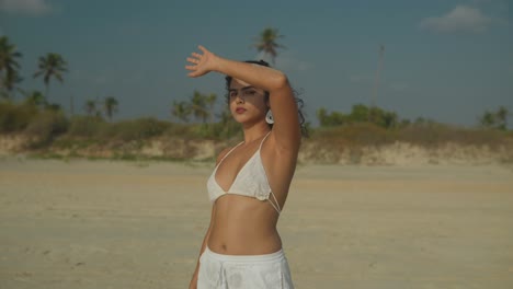 a young woman in white bikini and shorts stands on a beach, facing the camera