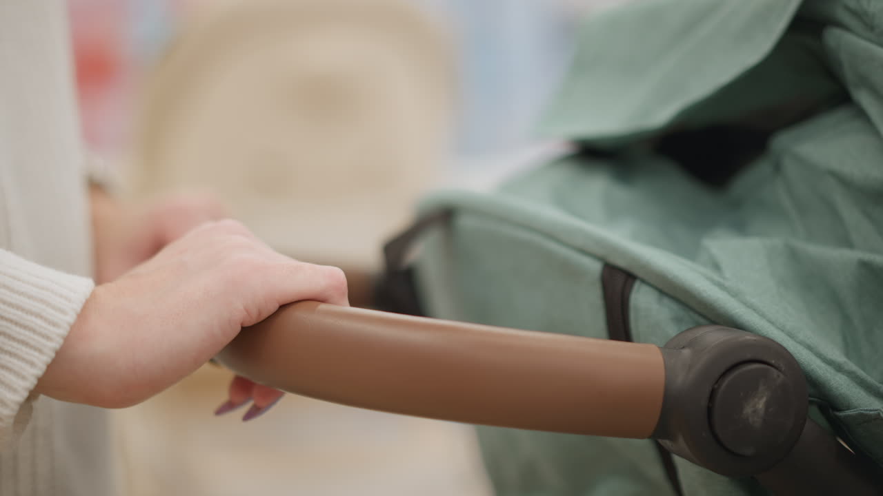 Hand view of mother holding stroller handle, pushing it gently forward and backward down well lit baby gear aisle in retail store while testing smooth maneuver and comfortable grip quality