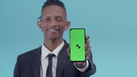office black man smiles and shows the screen of phone with a greenscreen on blue studio background