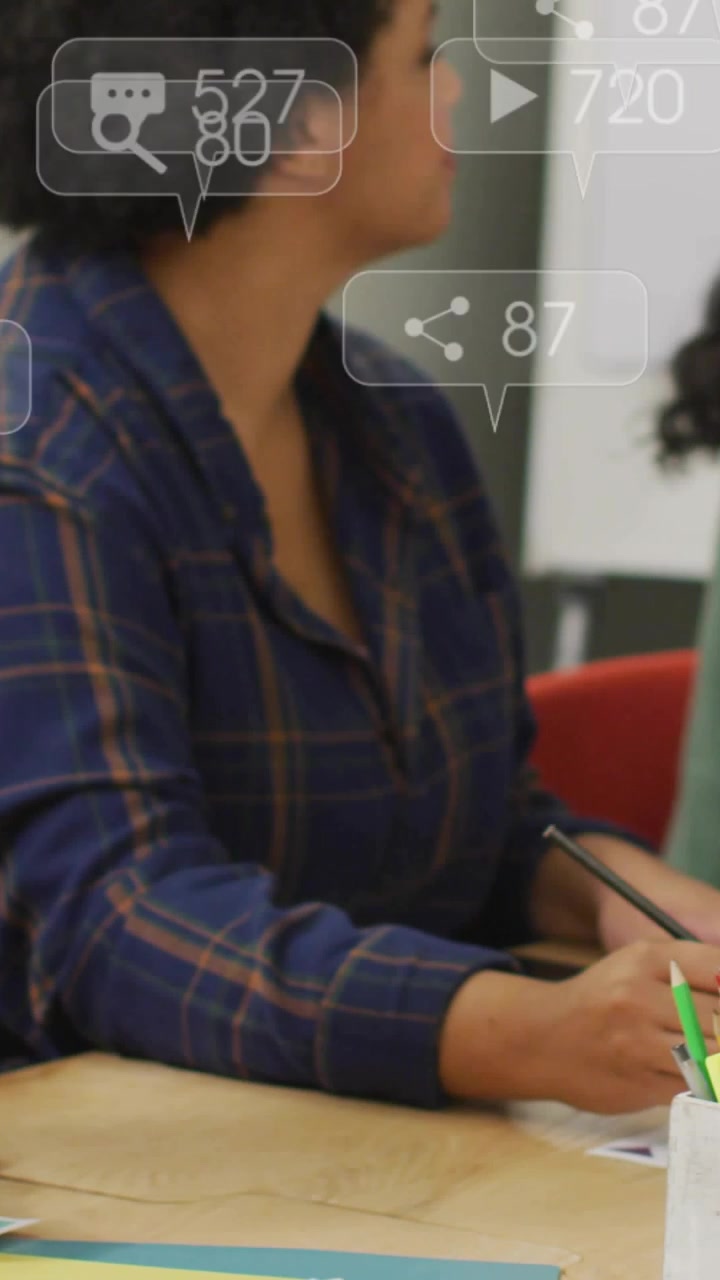 Video vertical: Mujer escribiendo mientras una presentadora de pie presenta una actualización de negocios, alertas flotando sobre un portátil.