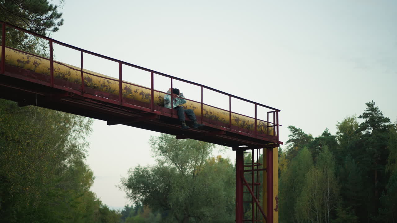 Viaggiatore seduto su un ponte sopraelevato di una condotta, con uno zaino accanto, che guarda la fitta chioma degli alberi, cielo di mezzogiorno, luce calda e soffusa, contrasto tra la ringhiera d'acciaio e la vegetazione lussureggiante, riposo tranquillo durante il viaggio
