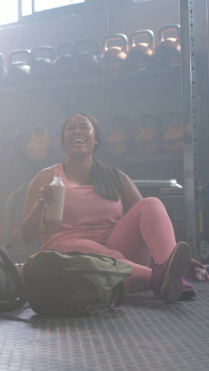 Video verticale: Donna che ride in rosa in palestra, controlla il polso, afferra uno shaker e si idrata vicino ai kettlebell