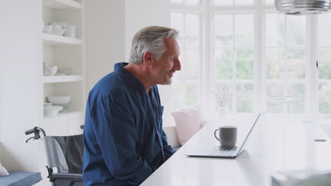 Älterer-Behinderter-Mann-Im-Rollstuhl-Zu-Hause,-Der-Einen-Videoanruf-Auf-Dem-Laptop-Auf-Der-Küchentheke-Führt