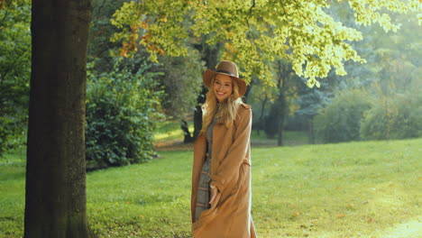 cheerful blonde young woman wearing hat and coat running and whirling in the park in autumn