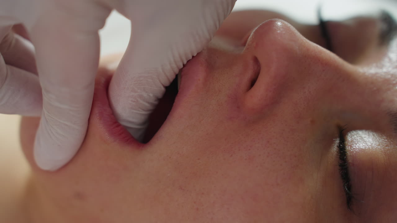 Close up of female client under clinical blanket, eyes closed and mouth open in relaxed manner as aesthetician wearing protective gloves dips fingertips into mouth and rubs jawline gently