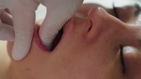 close up of female client under clinical blanket, eyes closed and mouth open in relaxed manner as aesthetician wearing protective gloves dips fingertips into mouth and rubs jawline gently