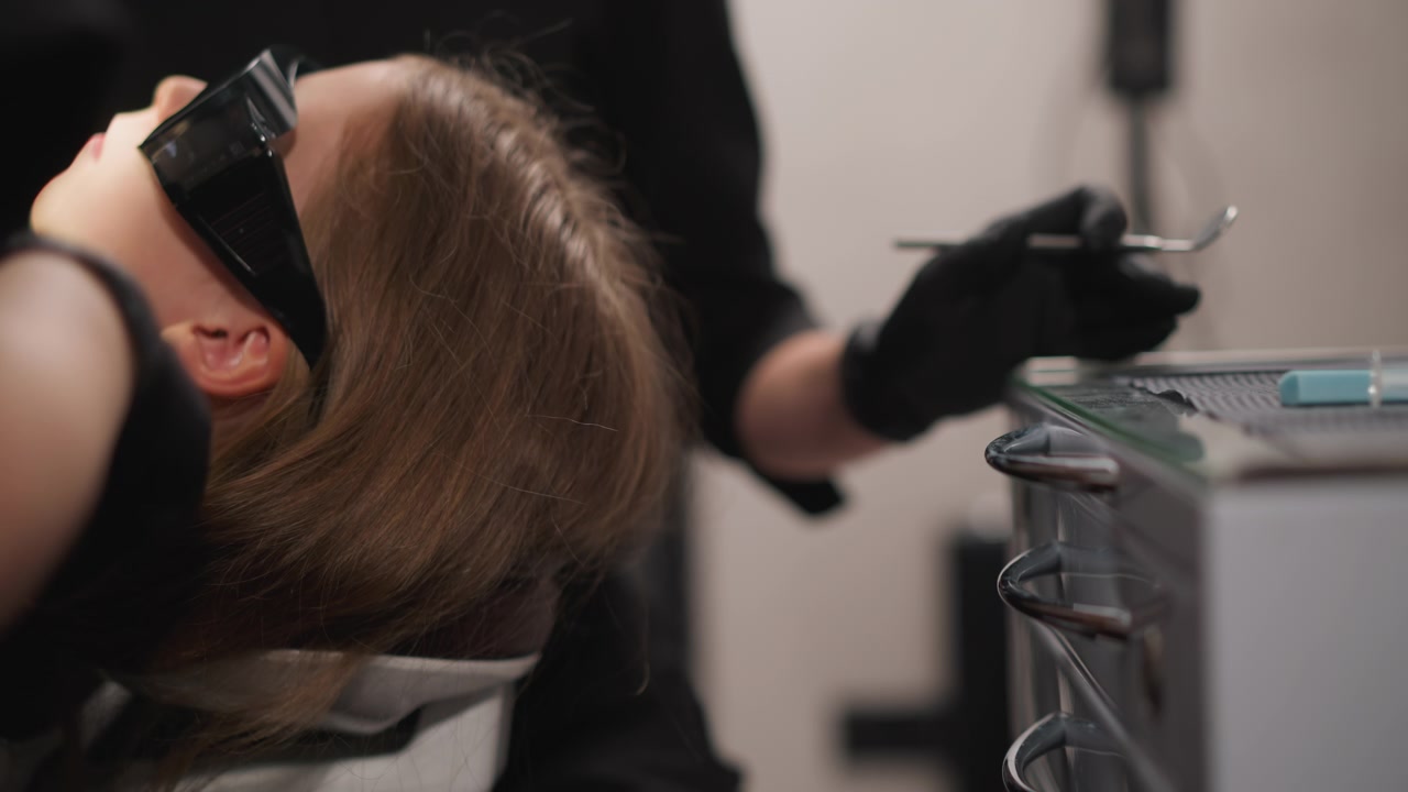 Primo piano di un professionista dentale che indossa guanti neri mentre prepara gli strumenti su un mobile metallico, assistendo un paziente adulto reclinato sullo sfondo. Strumenti e forniture dentali sono visibili.
