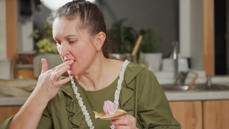 woman in green outfit holds swirl pink cupcake placed on paper while licking icing from finger and preparing to grab piping bag for next decorative coating in kitchen with baking supplies around