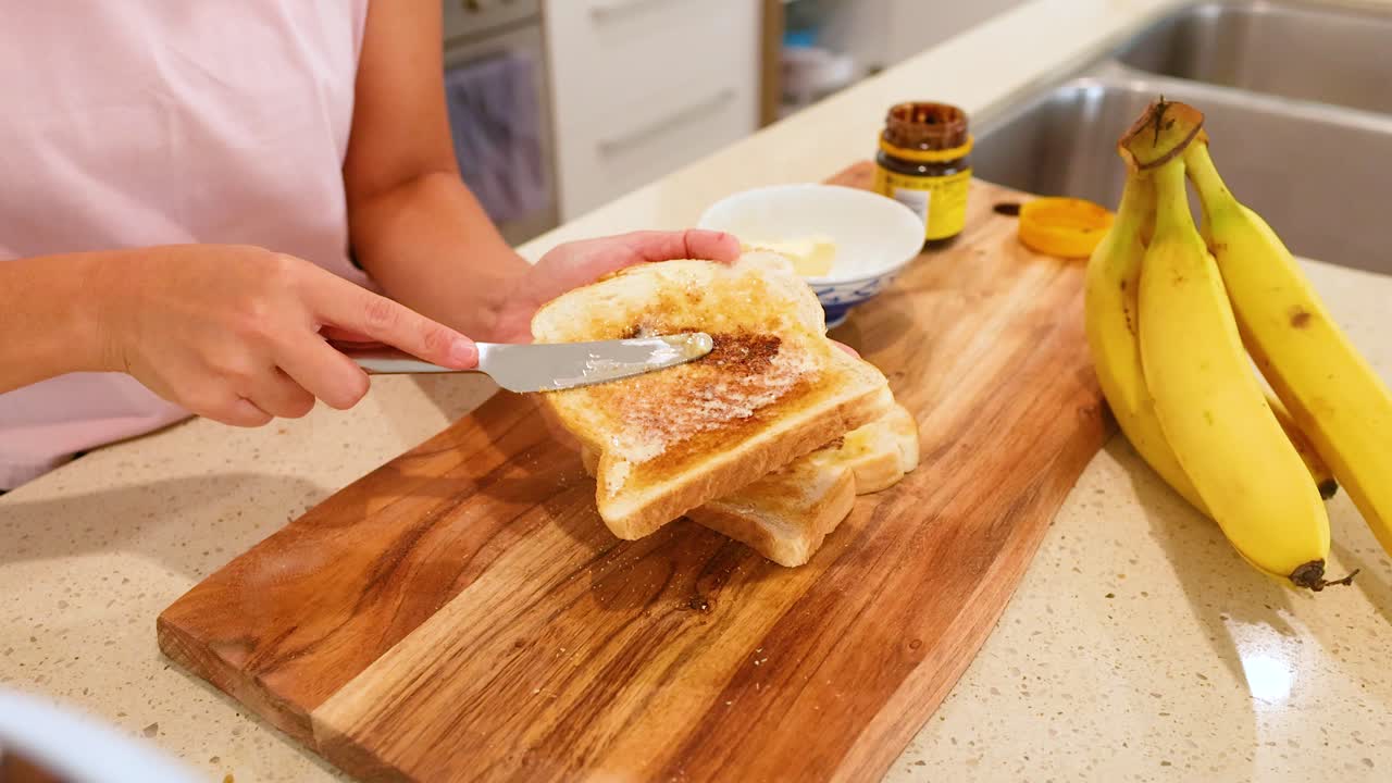 A person spreads Vegemite on buttered toast in a well-lit kitchen with bananas nearby