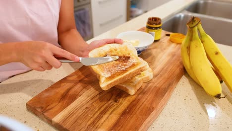 a person spreads vegemite on buttered toast in a well-lit kitchen with bananas nearby
