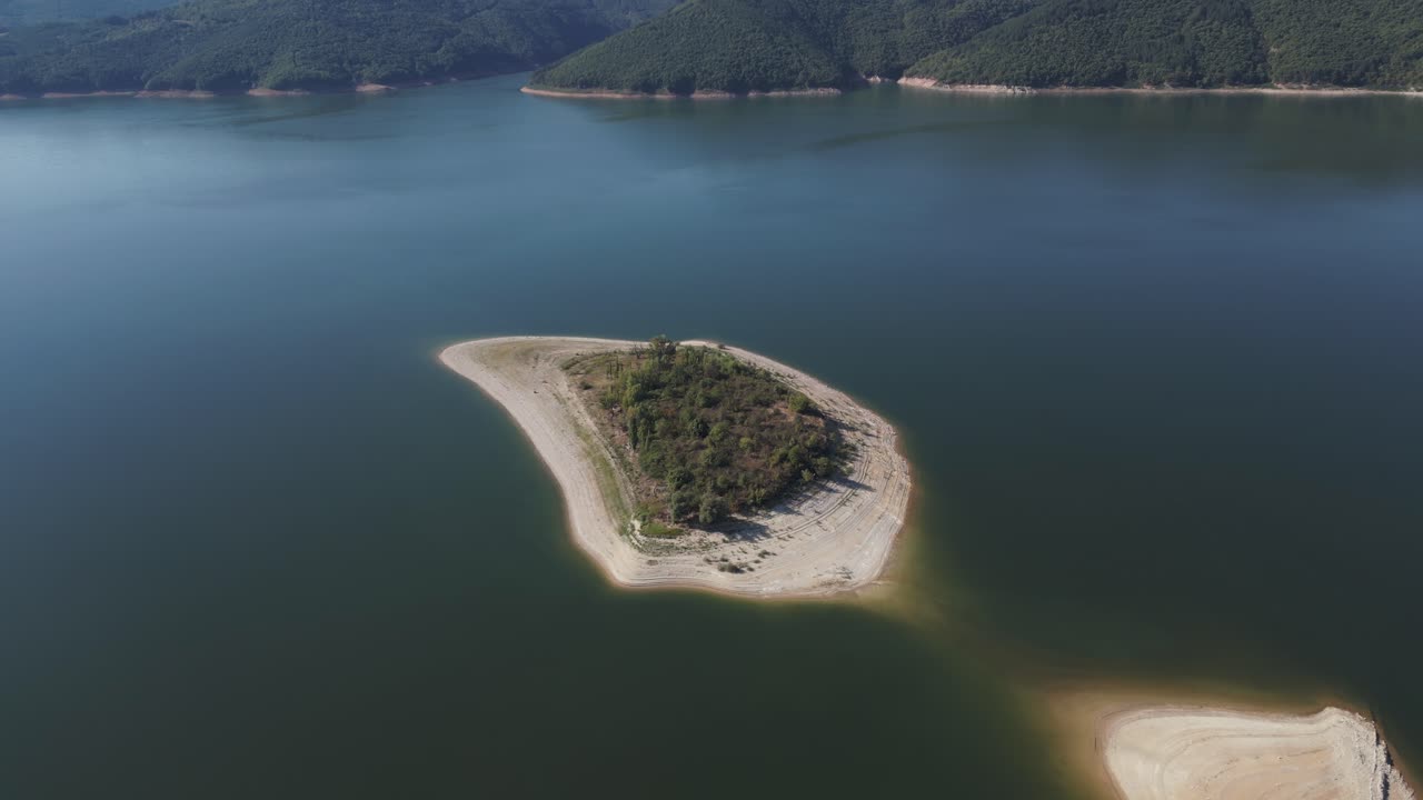 Aerial shot of a small, forested island surrounded by calm waters of a dam, highlighting natural beauty and serenity