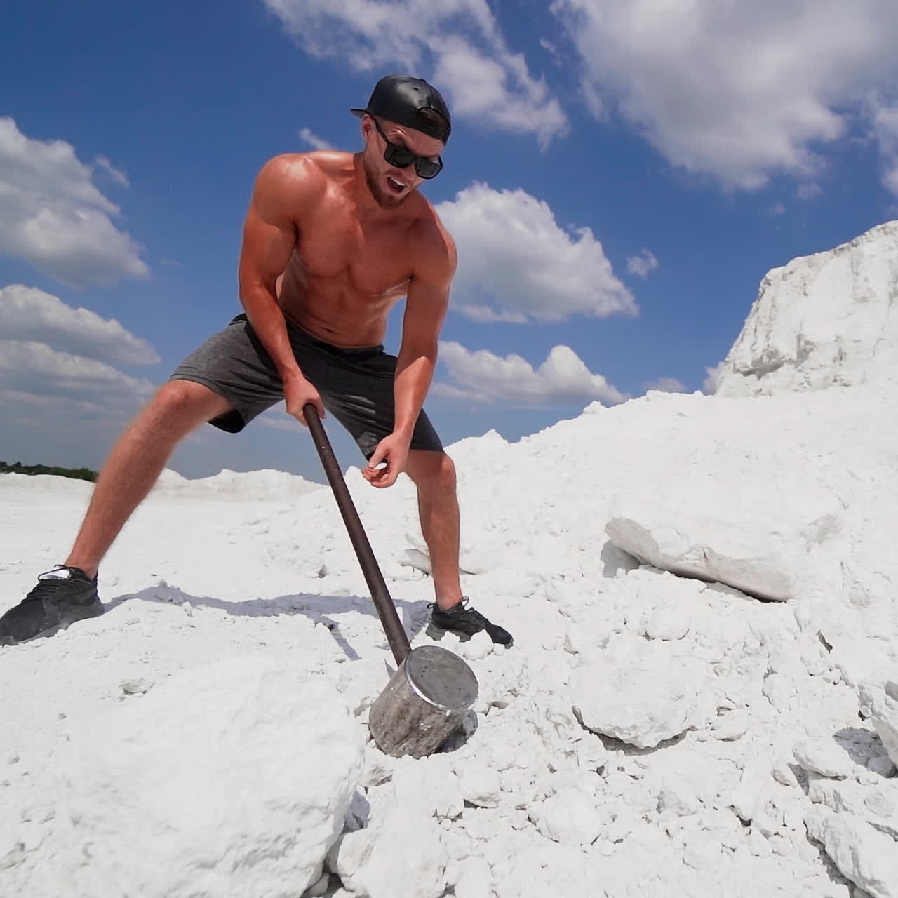 Muscular sportsman working in nature. Strong man with metal hammer breaking white rock on the mountain.