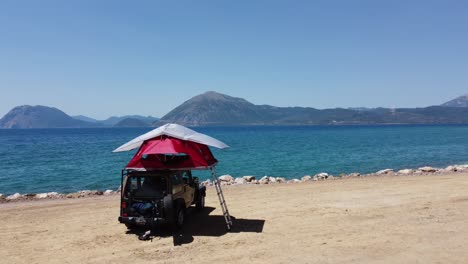 drone shot of a red roof top tent mounted on a 4x4 jeep at the beach with sea and mountains view on a sunny day