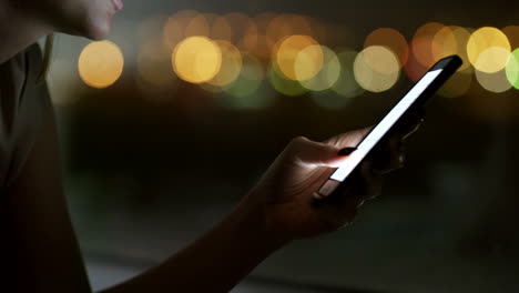 vista de cerca de una mujer escribiendo un mensaje en un smartphone en la oscuridad con luces desenfocadas en segundo plano