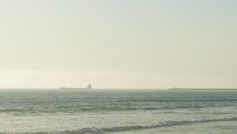 Cargo-Ship-Cruising-On-A-Foggy-Ocean-With-Waves-Coming-To-The-Beach-Shore-In-Figueira-da-Foz,-Portugal