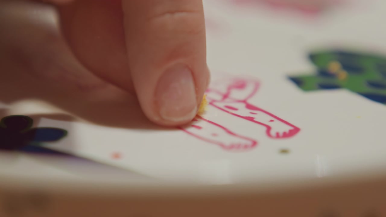Primo piano di una piccola briciola gialla su piatto bianco con disegni a cartone animato rosa e verdi. Una mano tocca delicatamente il bordo dell'inquadratura, indicando interazione senza disturbare la scena. Lo sfondo rimane.