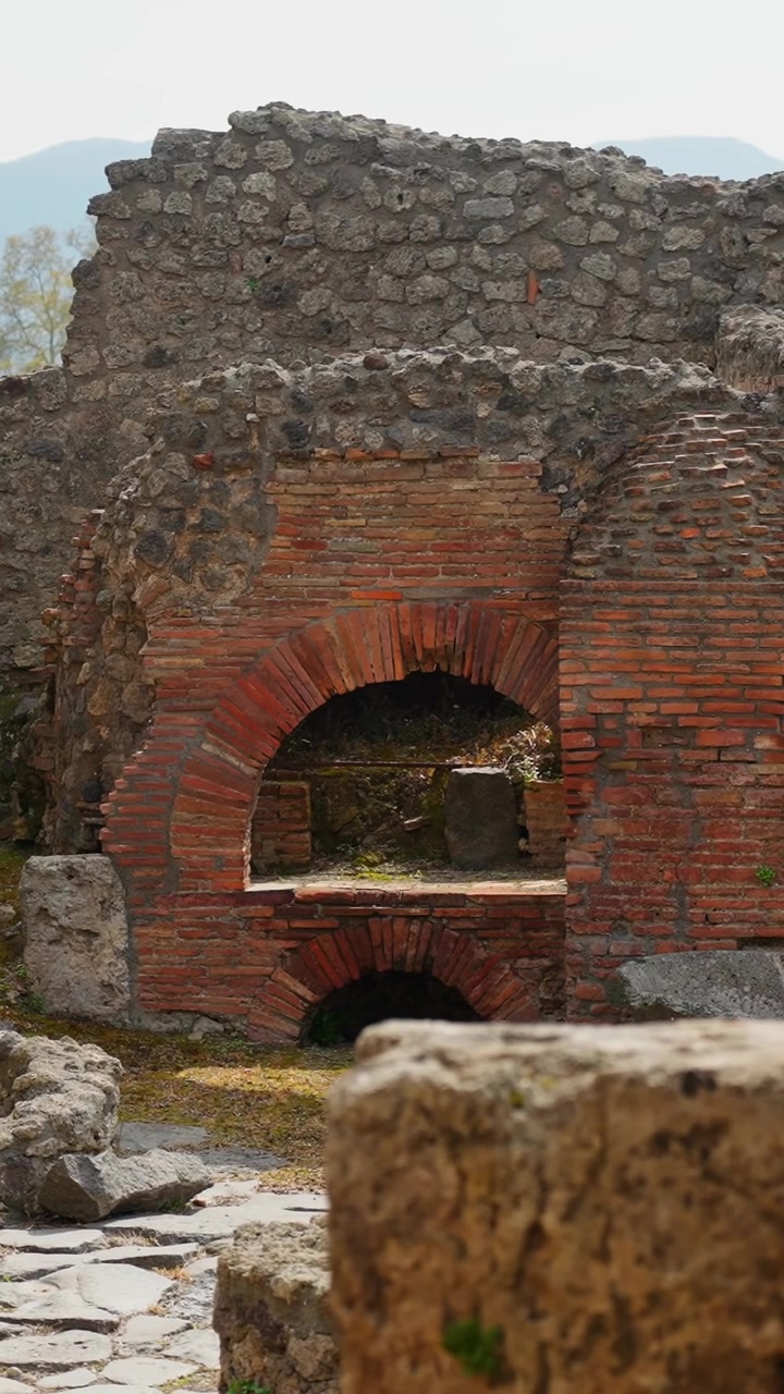 Vista verticale di un antico forno a mattoni romano o fornace nelle rovine di Pompei, con camere di combustione ad arco, muratura consumata dal tempo, muri in pietra e dettagli archeologici della produzione alimentare.