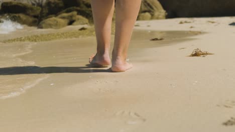 A-close-up-shot-of-the-heels-of-a-young-female-walking-leisurely-across-a-beach,-shallow-ocean-waves-washing-up-splashing-between-footsteps-as-she-continues-her-adventure