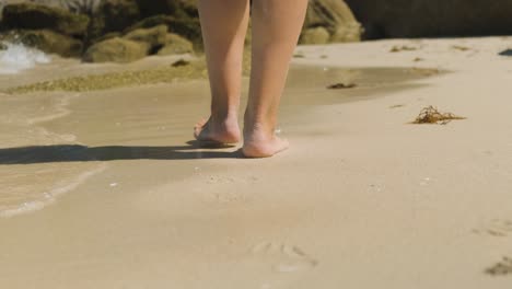 a close up shot of the heels of a young female walking leisurely across a beach, shallow ocean waves washing up splashing between footsteps as she continues her adventure