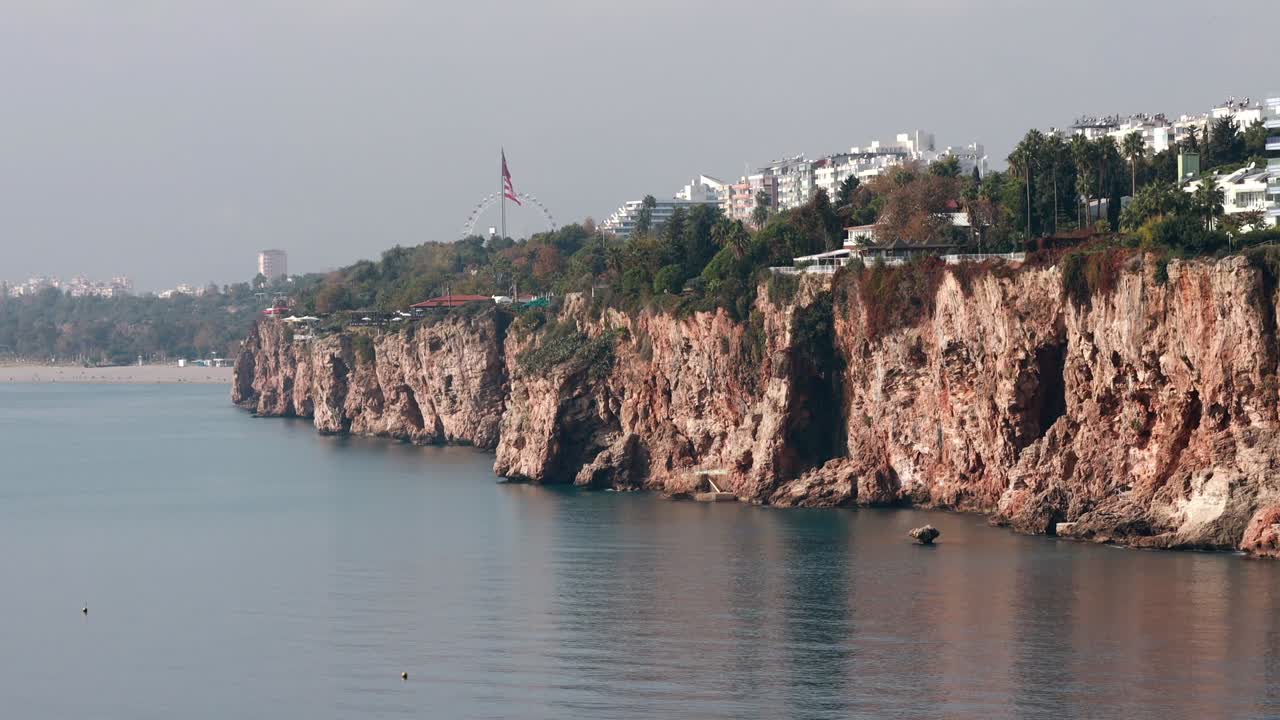 Drone view showing high coastal cliffs with white buildings ferris wheel and large flag above calm sea near Alanya in Antalya region with soft daylight and peaceful shoreline atmosphere