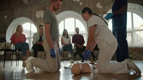 Close-up-shooting:-A-bearded-brunette-man-in-a-gray-T-shirt-along-with-a-professional-female-doctor-in-a-white-uniform-showing-first-aid-rules-with-a-mannequin-in-front-of-the-public-against-the-backdrop-of-brick-walls-and-large-windows