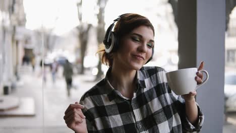 joyful, funny young girl listening to the music in headphones and dancing with a cup of coffee in hand. blurred street on the background