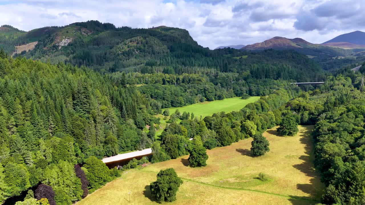 Drone glides above lush green forest, river, and mountains under bright daylight in Scottish Highlands