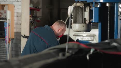 engineer in blue uniform working in mechanical shop with white car parked in background, industrial setting with tools and equipment, focused on repair task in organized workspace